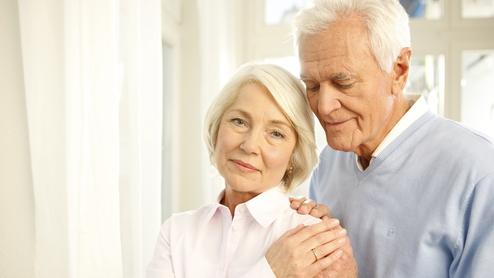 Ein älteres Paar steht nah beieinander, der Mann umarmt die Frau sanft, beide lächeln in eine ruhige Umgebung.